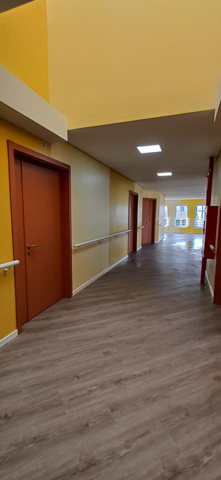 Corredor com portas em tom terracota e barras de apoio ao longo da parede.